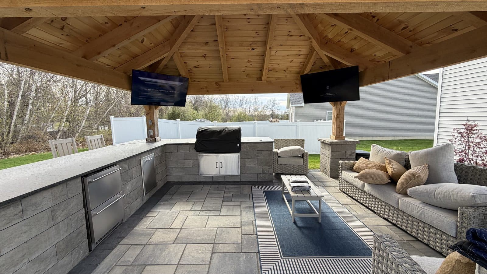 Two outdoor TVs mounted on the posts of a covered pergola with an outdoor kitchen and lounge seating